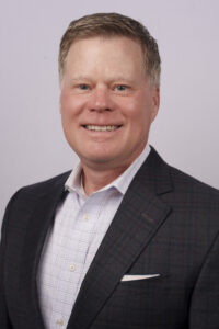 Professional head-and-shoulders portrait of a smiling man in a dark blazer and light checkered shirt against a pale purple background.
