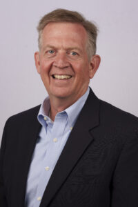 Smiling middle-aged man in a dark blazer and blue striped shirt, against a light gray background (corporate headshot).