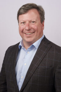 Portrait of a smiling man in a light blue shirt and brown checked blazer against a pale backdrop, a professional headshot.