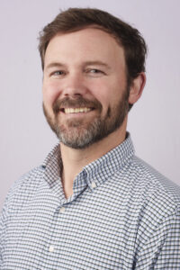Close-up portrait of a smiling man with a brown beard, wearing a blue-and-white checkered shirt.
