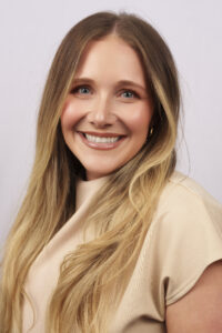 Close-up of a smiling woman with long blond hair and a beige top, looking at the camera.