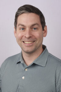 Close-up portrait of a smiling man with short dark hair and blue eyes, wearing a patterned button-down shirt against a light background.