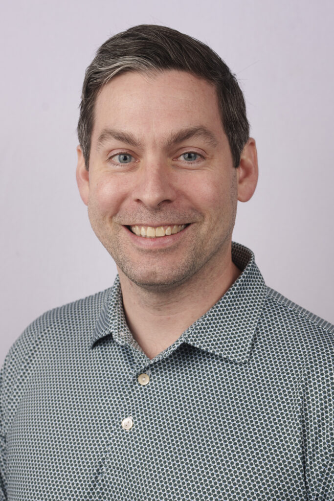 Close-up portrait of a smiling man with short dark hair and blue eyes, wearing a patterned button-down shirt against a light background.