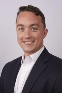 Smiling man in a dark blazer and white shirt, close-up portrait against a light gray background.