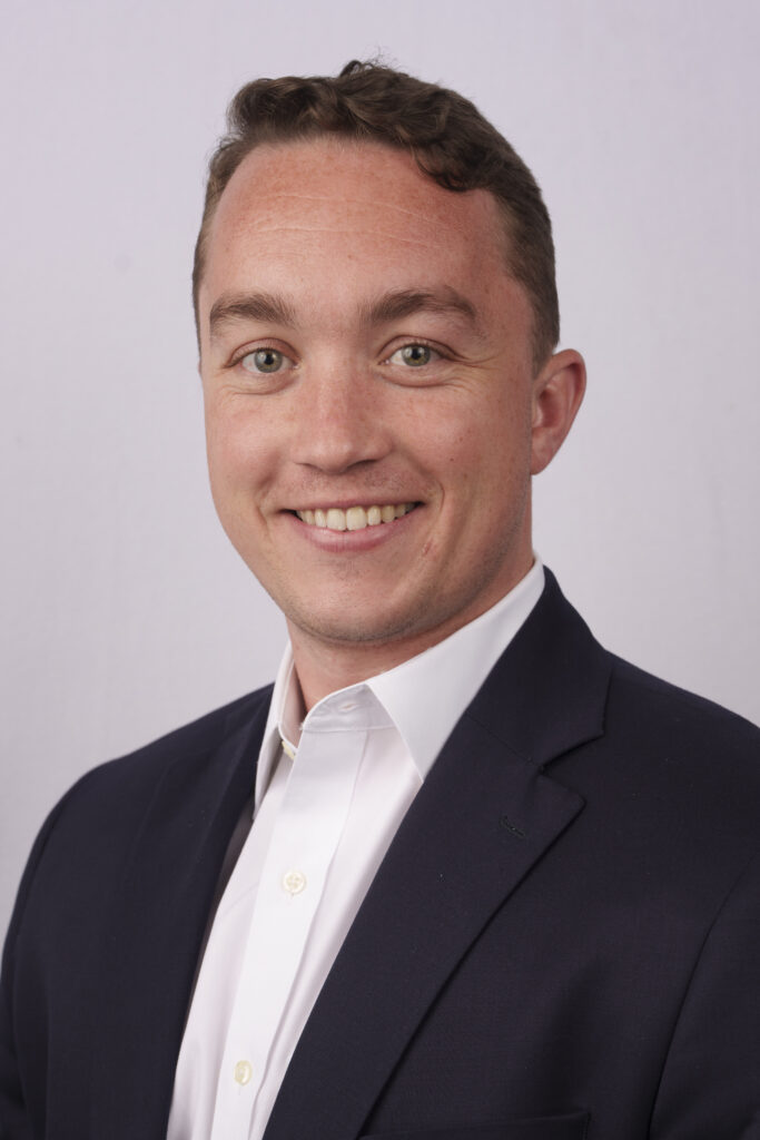 Smiling man in a dark blazer and white shirt, close-up portrait against a light gray background.