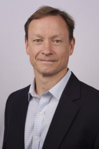 Professional headshot: man in a dark blazer and blue checkered shirt, smiling slightly, neutral background.