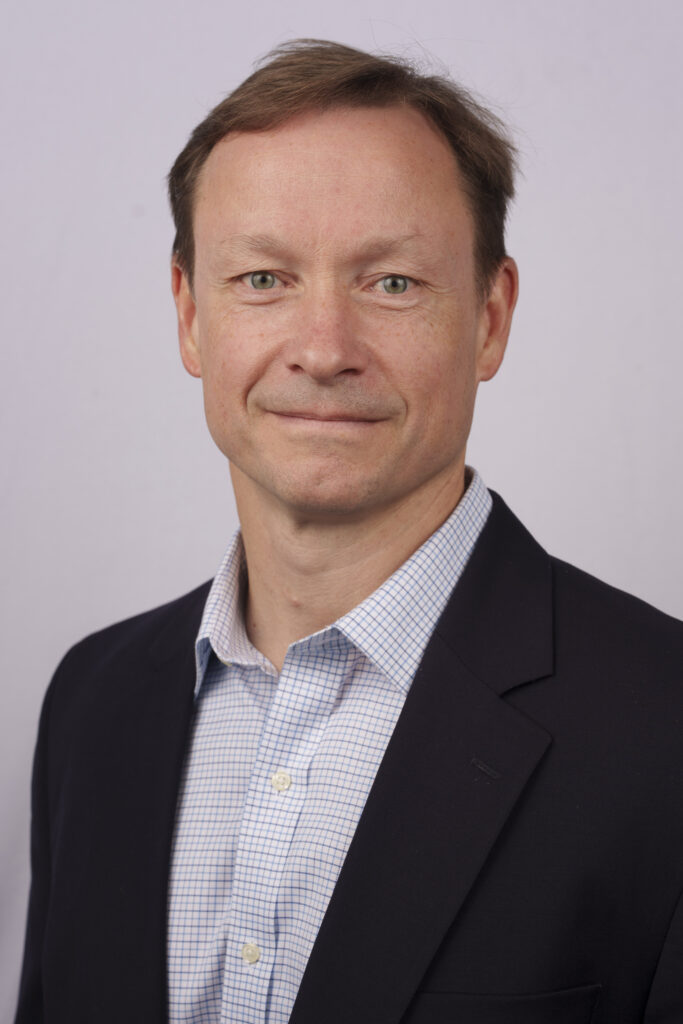 Professional headshot: man in a dark blazer and blue checkered shirt, smiling slightly, neutral background.
