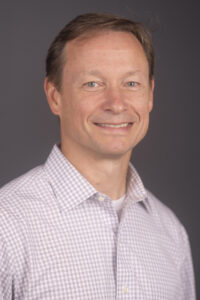 Head-and-shoulders portrait of a smiling man with short brown hair, wearing a light checkered shirt against a gray background.