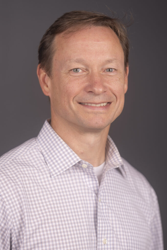 Head-and-shoulders portrait of a smiling man with short brown hair, wearing a light checkered shirt against a gray background.